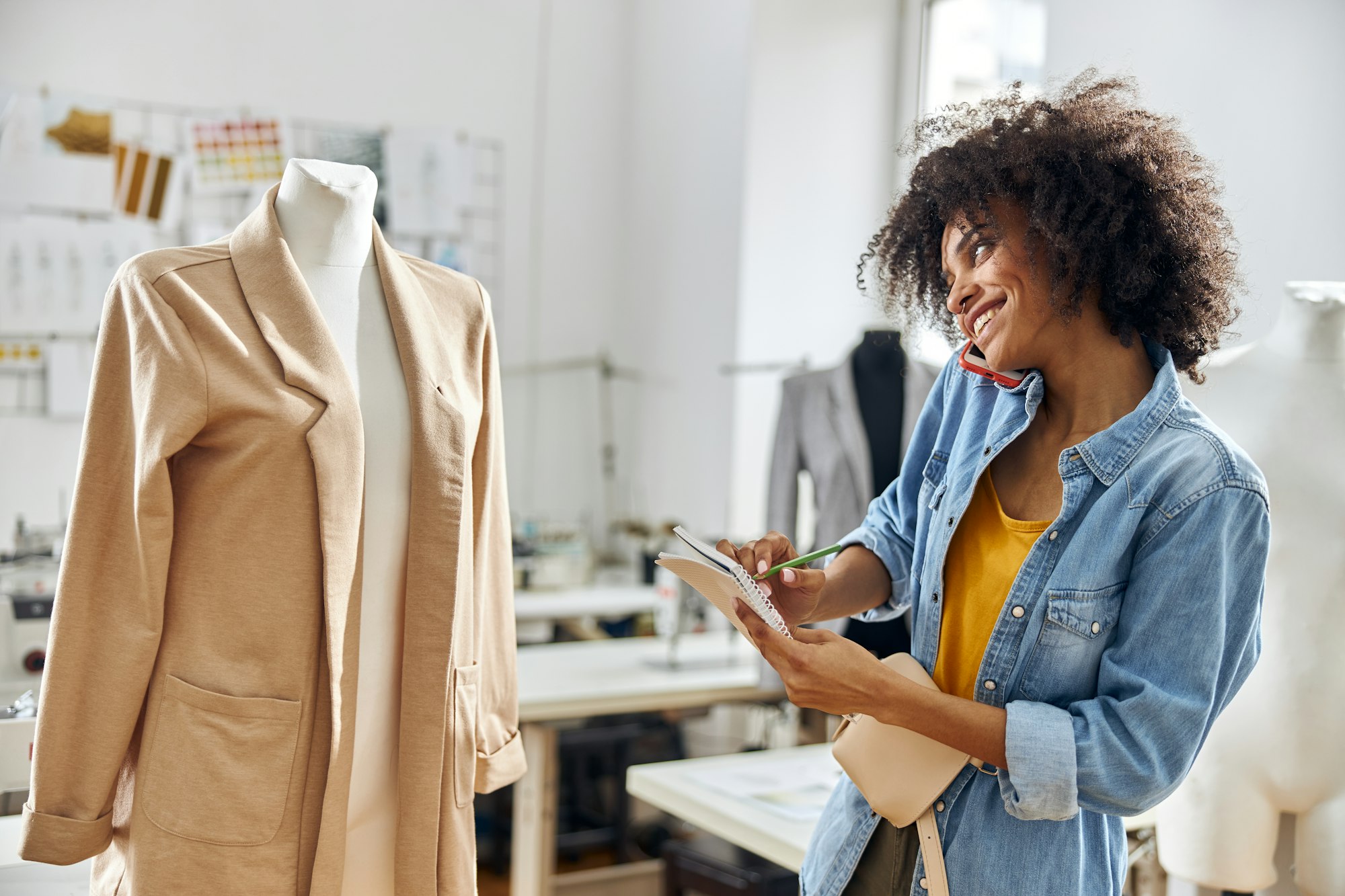 African-American woman talks on phone writing in notebook near mannequin in workshop